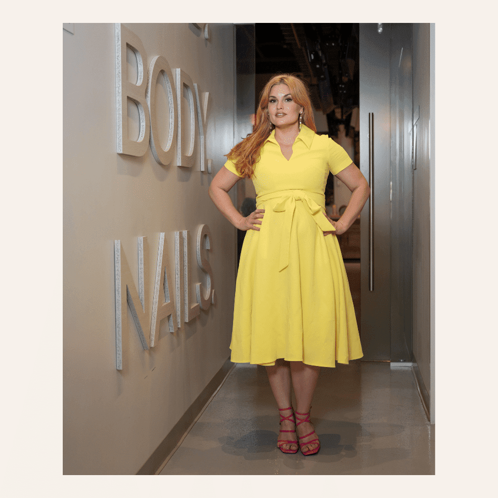 Model in yellow dress with pink heels stands confidently in hallway near wall signage.